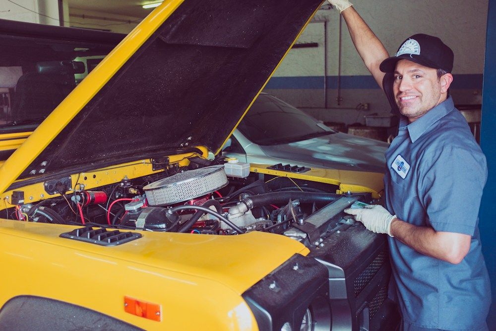 A smiling mechanic leaning on the open hood of a yellow car